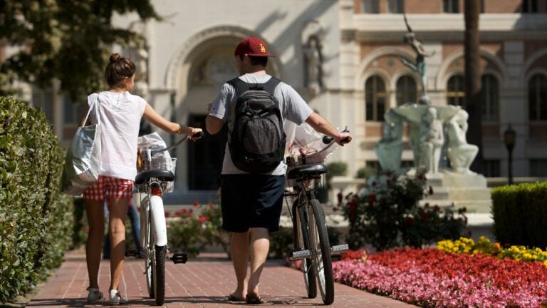 View from behind of two USC students pushing bicycles toward Doheny Library.