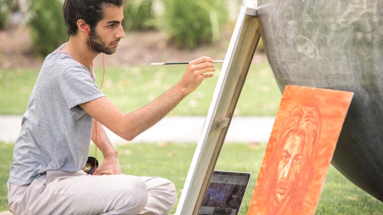 Student with headphones sits and paints outside on a canvas.