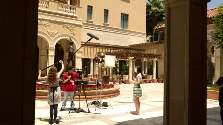 Students hold film equipment and record another student for a film project being shot near the USC School of Cinematic Arts.