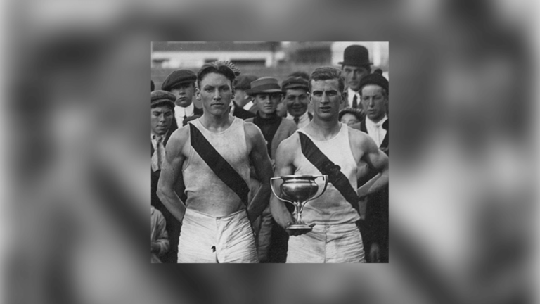 Black and white photo of 2 serious looking men, one with a trophy in their hand.