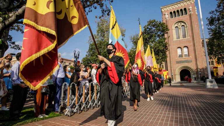 Students holding the flags of their college while walking during commencement.