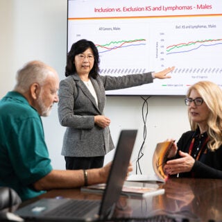 A woman explaining a graph being shown on a TV screen with another woman explaining something to an older man.
