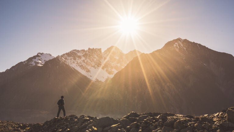 A person walks along a rock ridge with a landscape view of a mountain and sunset behind them.