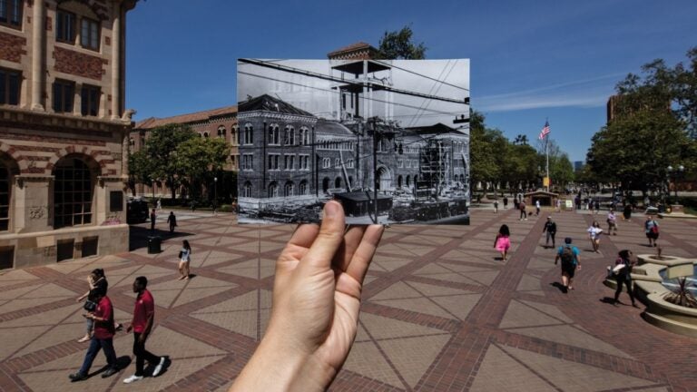 A hand holding out an old photo of campus in the exact area on campus in the modern day.