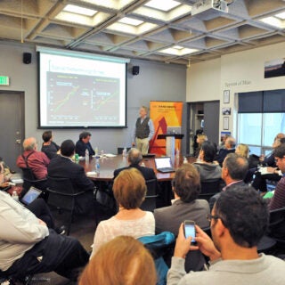 An audience sits in chairs formed in a circle around a main presenter who stands and speaks next to a presentation screen.