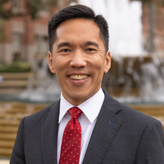 Beong-Soo Kim, USC president, smiling in a dark suit and red polka-dot tie, standing by a fountain on campus.