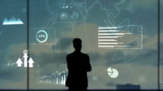 Silhouette of a person standing before large digital displays showing charts, graphs and maps.