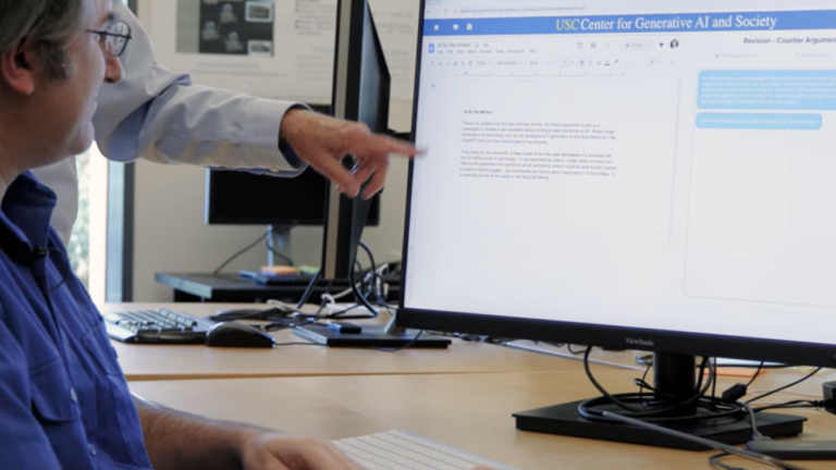 Two colleagues collaborate at a desk in an office as one points to a computer monitor displaying a writing document with revision comments.