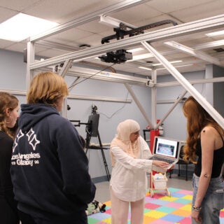 Instructor shows a laptop demonstration to students inside a biomechanics research lab with motion-tracking equipment.